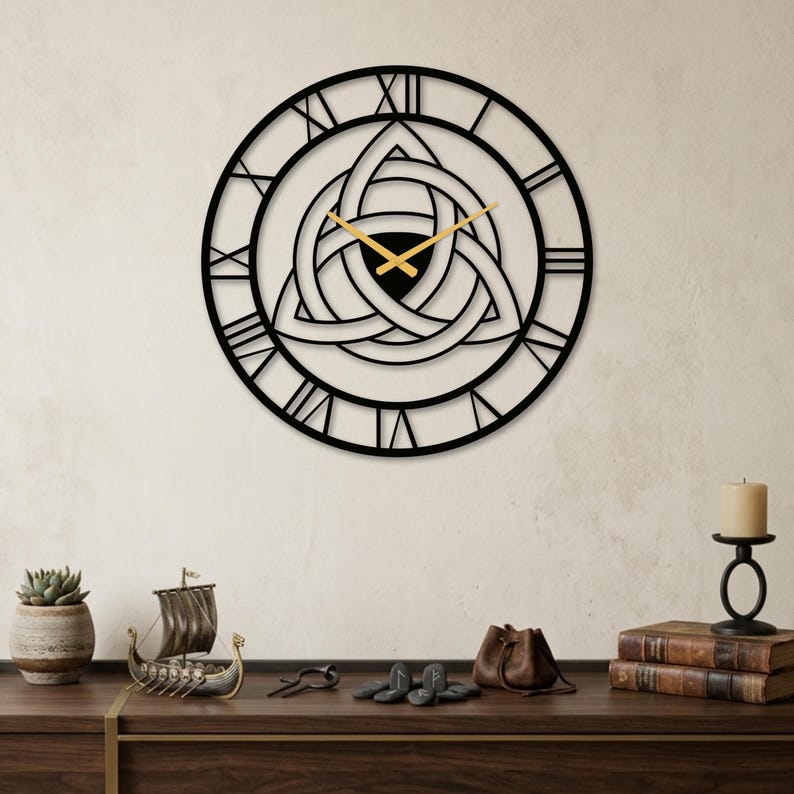 The large, black metal wall clock features a prominent central Triquetra (Trinity Knot) cut-out design and large Roman numerals. It has sleek modern gold hands and is mounted on a textured, light-colored wall. Below, on a rich, dark wood sideboard, a curated collection of Viking and Nordic themed decor is arranged. This metal clock and its associated decor create a unique, modern Nordic aesthetic perfect for a man cave, available on Etsy.
