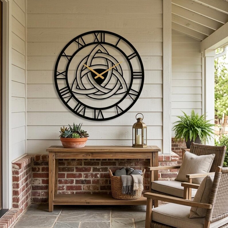 A large, black, laser-cut Metal Wall Clock with an intricate central Triquetra (Trinity Knot) and distinct Roman numerals is mounted on light-colored siding above a wooden console table on a covered Nordic porch. This striking piece of Viking Decor and Modern Wall Art features gold hands and is masterfully produced by Degary. It hangs above a rustic wooden table with a lantern and succulent, surrounded by wicker chairs, showing its unique style for unique Porch Decoration on Etsy.