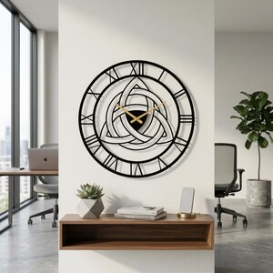 Degary Triquetra Metal Wall Clock with gold hands and Roman numerals, featuring an intricate Trinity Knot cut-out design and Roman numerals with gold hands, mounted on a white office wall. This large black metal wall clock is shown as a piece of Viking Office Decor and Modern Wall Art, positioned above a wooden floating shelf with a succulent and books. It's displayed in a modern office context suitable for an Etsy listing, with workstations, chairs, and windows with a city view.