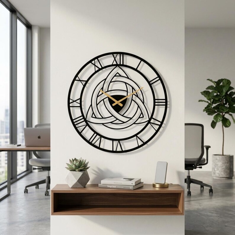 Degary Triquetra Metal Wall Clock with gold hands and Roman numerals, featuring an intricate Trinity Knot cut-out design and Roman numerals with gold hands, mounted on a white office wall. This large black metal wall clock is shown as a piece of Viking Office Decor and Modern Wall Art, positioned above a wooden floating shelf with a succulent and books. It's displayed in a modern office context suitable for an Etsy listing, with workstations, chairs, and windows with a city view.