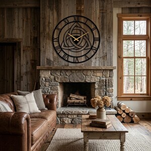 A large, black, laser-cut Metal Wall Clock with an intricate central Triquetra (Trinity Knot) design and distinctive Roman numerals is prominently displayed on a rustic wood-plank wall above a stone fireplace in a cozy cabin living room. This detailed piece of Modern Wall Art, masterfully created by Degary, serves as a striking centerpiece for a Nordic and Viking-inspired home.