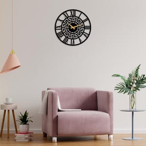 Large Meander metal wall clock featuring an ancient Greek key pattern, modern black steel wall art decor hanging on a minimalist wall behind a pink armchair in a stylish living room.
