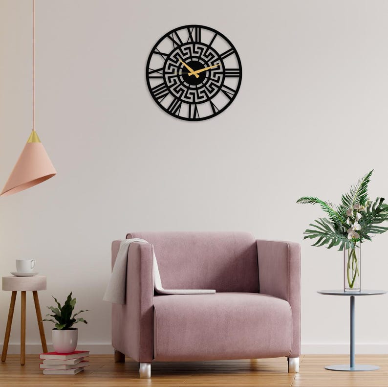 Large Meander metal wall clock featuring an ancient Greek key pattern, modern black steel wall art decor hanging on a minimalist wall behind a pink armchair in a stylish living room.