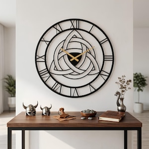 Large black metal Triquetra wall clock by Degary hanging on a white wall. This 2mm thick steel laser-cut clock features Roman numerals, a central Celtic Trinity knot design, and gold hands. Displayed above a wooden console table styled with rustic Viking decor, including miniature horned helmets, Thor's hammer, and runestones.
