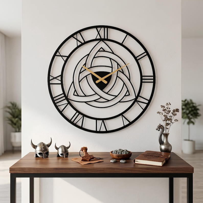 Large black metal Triquetra wall clock by Degary hanging on a white wall. This 2mm thick steel laser-cut clock features Roman numerals, a central Celtic Trinity knot design, and gold hands. Displayed above a wooden console table styled with rustic Viking decor, including miniature horned helmets, Thor's hammer, and runestones.