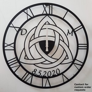 A personalized matte black Metal Wall Clock by Degary, featuring a laser-cut Triquetra (Trinity Knot) design customized with the initials 'D' and 'M' alongside an anniversary date '8.5.2020'. The clock displays classic Roman numerals and sleek silver hands against a light background, highlighting its use as a Personalized Wedding Gift, Anniversary Gift, Viking Decor, and customizable Modern Wall Art on Etsy.