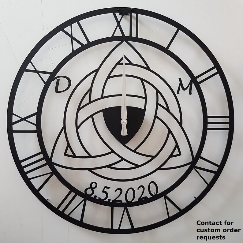 A personalized matte black Metal Wall Clock by Degary, featuring a laser-cut Triquetra (Trinity Knot) design customized with the initials 'D' and 'M' alongside an anniversary date '8.5.2020'. The clock displays classic Roman numerals and sleek silver hands against a light background, highlighting its use as a Personalized Wedding Gift, Anniversary Gift, Viking Decor, and customizable Modern Wall Art on Etsy.