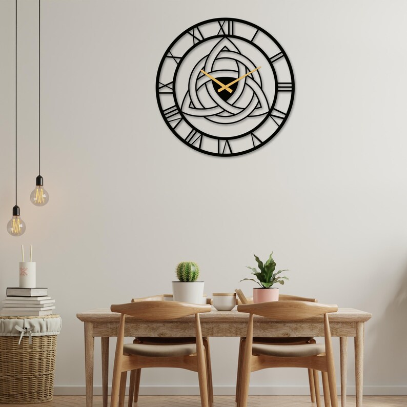 Metal wall clock with an intricate Triquetra and Trinity Knot center, Roman numerals, and gold hands, masterfully produced by Degary. This piece of Modern Wall Art and Viking Decor is centrally mounted in a Nordic Dining Room Decoration, hanging over a light-wood dining table with two chairs. The table is styled with potted succulents, and the scene includes minimalist pendant lights and a woven basket with books. Ideal for creating a curated, Scandinavian atmosphere.