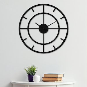 Puede incluir: Un reloj de pared negro y circular con un diseño minimalista. El reloj presenta una esfera redonda con un dial central y manecillas que indican la hora. El reloj está montado en una pared blanca, con una pequeña mesa debajo.