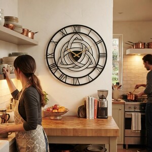 A large, black, laser-cut Metal Wall Clock by Degary, featuring an intricate central Triquetra (Trinity Knot) design, Roman numerals, and sleek gold hands, is mounted on a white wall in a warm, lively kitchen setting. The cozy lifestyle scene highlights a beautiful Nordic Kıtchen Decoration where a woman in an apron stands, while a man cooks at a stove in the background. This elegant piece of Modern Wall Art and Viking Decor complements a rustic or contemporary Scandinavian home aesthetic.