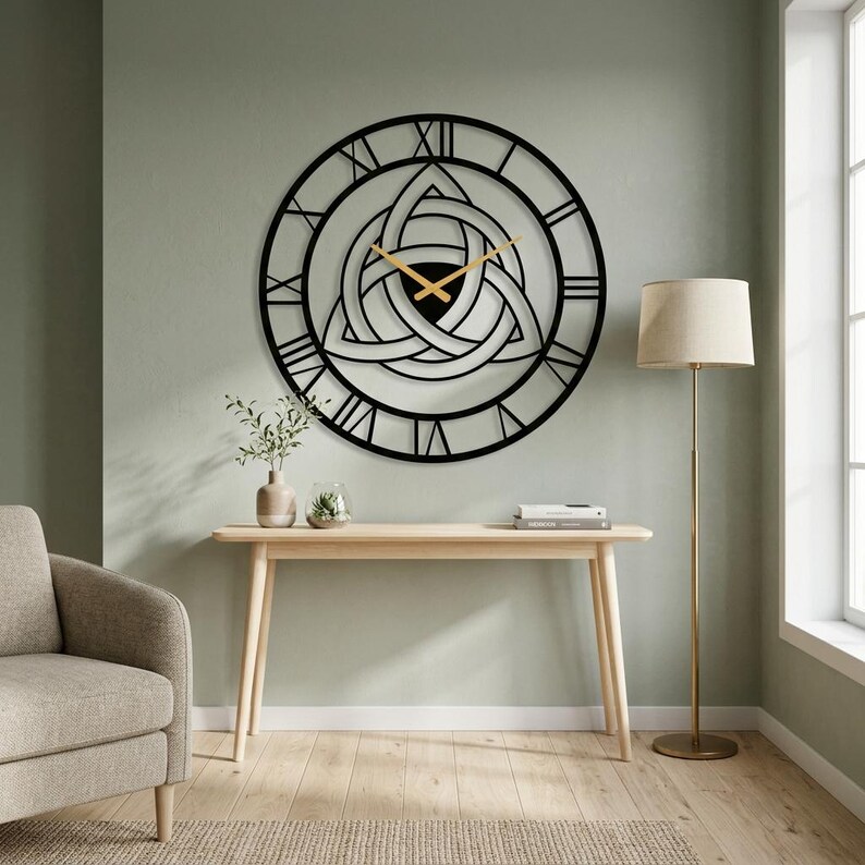 Large black Metal Wall Clock featuring a central Triquetra Celtic knot design and Roman numerals, Masterfully Produced by Degary. The stunning piece of Viking Wall Art and Nordic Home Decoration hangs above a light wood console table in a modern living room setting with a green wall. The scene includes an armchair, plants, and a floor lamp, perfect for any curate room and available on Etsy.
