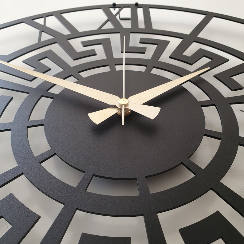 Close-up detail of a black metal wall clock with an intricate Greek key pattern and contrasting gold hands, modern minimalist decor element.