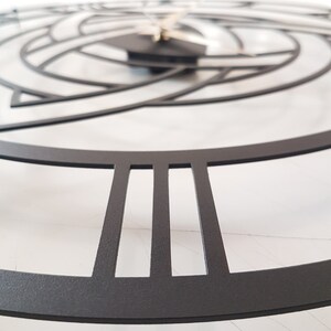 A detailed close-up macro shot of a matte black Metal Wall Clock, featuring a sharp focus on the precise laser-cut Roman numerals and the premium textured surface. The shallow depth of field elegantly blurs the background, softly revealing the intricate central Triquetra Trinity Knot design and clock hands. This perspective highlights the high-quality craftsmanship of the Modern Wall Art and Viking Decor, resting on a clean white marble background. Ideal for showcasing product textures on Etsy