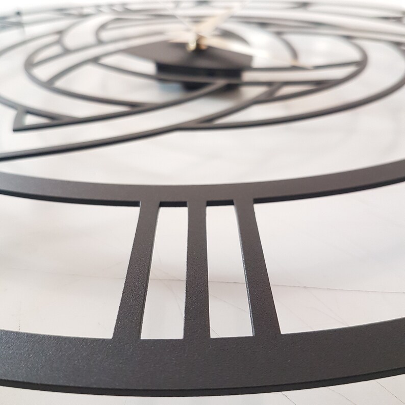 A detailed close-up macro shot of a matte black Metal Wall Clock, featuring a sharp focus on the precise laser-cut Roman numerals and the premium textured surface. The shallow depth of field elegantly blurs the background, softly revealing the intricate central Triquetra Trinity Knot design and clock hands. This perspective highlights the high-quality craftsmanship of the Modern Wall Art and Viking Decor, resting on a clean white marble background. Ideal for showcasing product textures on Etsy