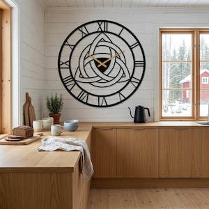 A large laser-cut Metal Wall Clock by Degary. This striking timepiece features elegant Roman numerals on the outer dial and an intricate, central Triquetra (Trinity Knot) design with sleek gold hands. The clock is prominently mounted on a white wood-paneled wall above an L-shaped light oak wood kitchen counter. The Scandinavian Kıtchen Decoration setting includes a loaf of bread, cutting boards, ceramic mugs, a black kettle, and a potted plant, with windows overlooking a snowy landscape.
