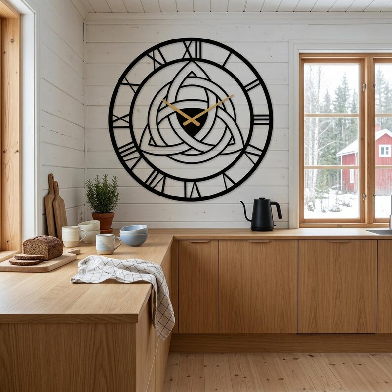 A large laser-cut Metal Wall Clock by Degary. This striking timepiece features elegant Roman numerals on the outer dial and an intricate, central Triquetra (Trinity Knot) design with sleek gold hands. The clock is prominently mounted on a white wood-paneled wall above an L-shaped light oak wood kitchen counter. The Scandinavian Kıtchen Decoration setting includes a loaf of bread, cutting boards, ceramic mugs, a black kettle, and a potted plant, with windows overlooking a snowy landscape.