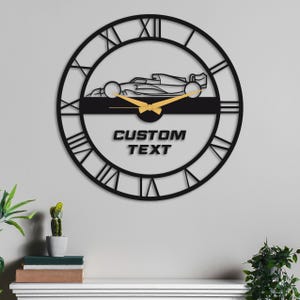 May include: Black metal wall clock with Roman numeral markings and a silhouette of a race car. The clock has gold-colored hands and a space for custom text that reads "CUSTOM TEXT".