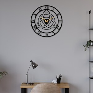 A Metal Wall Clock, featuring an intricate central Triquetra design, Roman numerals, and elegant gold hands. The clock is mounted on a minimalist light gray wall in a modern workspace, perfectly illustrating Nordic Home Office Decoration. Below, a wooden desk is styled with a metallic silver desk lamp, accompanied by a beige office chair. A black shelving unit with a trailing green plant is visible on the right. This Modern Wall Art and Viking Decor is ideal for contemporary workspaces.