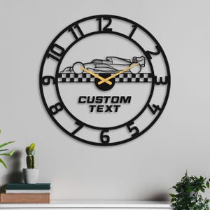 May include: Black metal wall clock featuring a race car design. The clock has a circular frame with cut-out numbers and hands. The words "CUSTOM TEXT" are below the race car image. The clock is a decorative item.