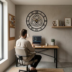 A large, black, laser-cut Metal Wall Clock by Degary, showcasing an intricate central Triquetra (Trinity Knot) design and distinctive Roman numerals, hangs prominently on a textured grey concrete-look wall in a Modern Office workspace. A man is seated at a wooden desk, working on a laptop, with minimalist Nordic Decoration elements, illustrating this unique piece as Viking Decor and curated Modern Office Wall Art for curated Home Decor