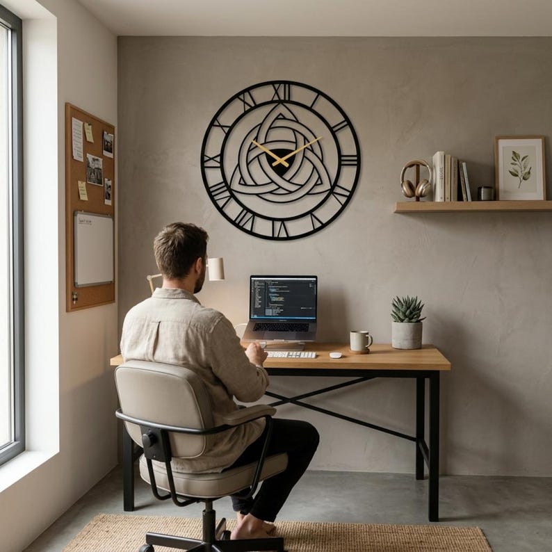 A large, black, laser-cut Metal Wall Clock by Degary, showcasing an intricate central Triquetra (Trinity Knot) design and distinctive Roman numerals, hangs prominently on a textured grey concrete-look wall in a Modern Office workspace. A man is seated at a wooden desk, working on a laptop, with minimalist Nordic Decoration elements, illustrating this unique piece as Viking Decor and curated Modern Office Wall Art for curated Home Decor