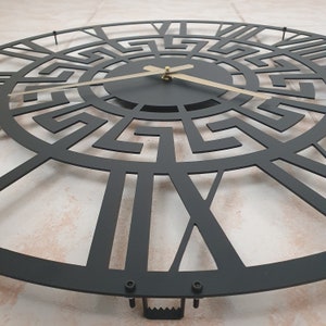 Close-up photo of hanger on the back side of a black metal wall clock with an intricate Greek key pattern and contrasting gold hands.