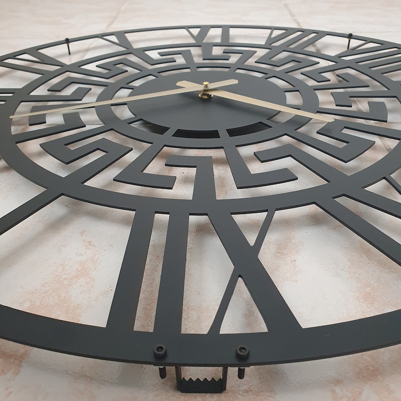 Close-up photo of hanger on the back side of a black metal wall clock with an intricate Greek key pattern and contrasting gold hands.