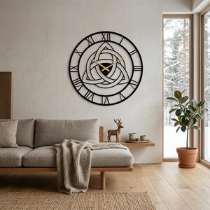 A modern living room featuring a large, black, laser-cut Metal Wall Clock with Roman numerals and a central Triquetra Trinity Knot design. This striking piece of Modern Wall Art and Viking Decor is mounted above a Nordic-style light-wood sofa with a beige knitted blanket. The room includes a jute rug, a potted rubber plant, a decorative wooden deer figurine, and a large window with a scenic snowy view, illustrating high-end Scandi and Nordic Living Room Decoration.