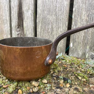May include: A copper saucepan with a dark brown handle. The saucepan has a hammered finish and a small circular stamp on the bottom that reads "Cuivre Pur."