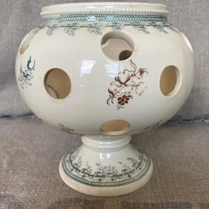 May include: A cream-coloured ceramic bulb vase with a spherical body and a pedestal base. The vase features multiple circular openings for flower stems, decorated with green patterned borders and brown floral illustrations on the exterior surface.