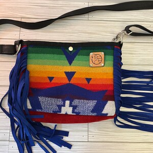 May include: A colorful crossbody bag with a southwestern design. The bag features a rainbow pattern with blue fringe and a black strap. A leather patch with a cactus logo is on the front.