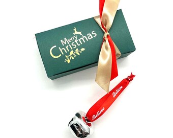 May include: A green gift box with gold text that reads "Merry Christmas" and a reindeer graphic. The box is tied with red and gold satin ribbon. A silver bell with a red ribbon that says "Believe" is also in the image.