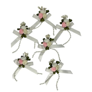 Puede incluir: Seis lazos de satén blanco con flores de tela rosa y blanca y detalles de perlas. Los lazos están atados con un lazo y tienen una larga cadena de perlas que cuelga del centro.