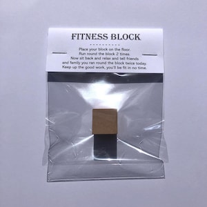 Könnte beinhalten: Ein kleiner brauner Holzblock in einem durchsichtigen Plastikbeutel mit einem weißen Etikett, auf dem "FITNESS BLOCK" steht und Anweisungen zum "Legen Sie Ihren Block auf den Boden. Laufen Sie zweimal um den Block. Setzen Sie sich jetzt zurück, entspannen Sie sich und erzählen Sie Ihren Freunden und Ihrer Familie, dass Sie heute zweimal um den Block gelaufen sind. Machen Sie weiter so, Sie werden in kürzester Zeit fit sein."