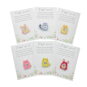 Op de afbeelding: Zes fidget-speeltjes op kaarten. Elke kaart heeft de tekst "Fidget friend" en een gedicht. De fidget-speeltjes hebben de vorm van dieren met een draaiend centrum. De dieren zijn een beer, een wasbeer, een beer, een kat en een konijn.