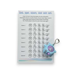 May include: A score card for the game Rock, Paper, Scissors, Shoot, with a light blue mini game. The card has game results and instructions. The mini game is light blue with a silver keychain.