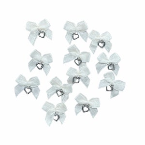 May include: A group of white satin bows, each featuring a small silver heart charm. The bows are set against a white backdrop, giving a delicate and feminine appearance. Suitable for crafting or decorating various items.