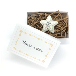 Puede incluir: Una caja de regalo blanca con un broche en forma de estrella en su interior, sobre papel marrón. El broche es verde claro y blanco con una joya en forma de flor. La tapa de la caja tiene el texto "You're a star" con borde de estrellas.