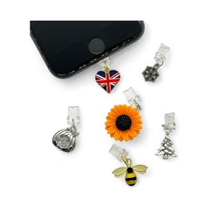 May include: A collection of phone charms with various designs, including a Union Jack heart, a sunflower, a bee, a snowflake, a pumpkin, and a Christmas tree. The charms are attached to clear phone plug adapters.