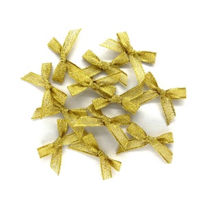 Puede incluir: Un montón de pequeños lazos dorados hechos de cinta brillante. Los lazos están esparcidos sobre un fondo blanco.