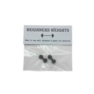 Op de afbeelding: Twee kleine, donkergrijze halter-oorbellen worden getoond in een doorzichtige plastic zak. De zak is bevestigd aan een witte kaart met de tekst "BEGINNERS WEIGHTS" en "Easy to use, well balanced & great for workouts."