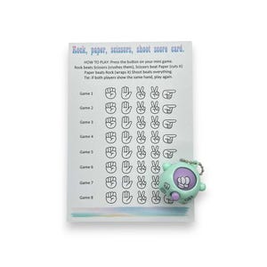 May include: A score card for the game "Rock, Paper, Scissors, Shoot" with instructions and game results. A small, mint green game device with a purple button is attached to the card. The text on the card reads "Rock, paper, scissors, shoot score card."