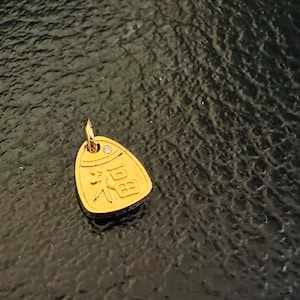 May include: A gold-toned pendant shaped like a rounded triangle, featuring a small embedded clear stone at the top and the Chinese character for good fortune, Fu, embossed in the center, complete with a small circular jump ring for attachment.