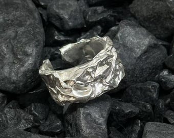 May include: A silver ring with a textured, crumpled appearance. The ring is set against a backdrop of dark gray rocks, creating a contrast between the smooth metal and the rough stone.