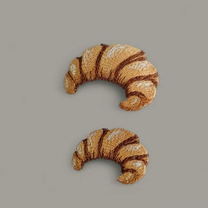 May include: Two embroidered felt patches shaped like croissants. The larger croissant is brown with a white swirl design. The smaller croissant is brown with a white swirl design.