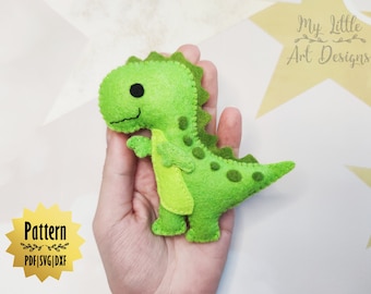 May include: A green felt dinosaur toy with black eyes and a stitched smile. The dinosaur is being held in a hand. The text "My Little Art Designs" is visible in the background.