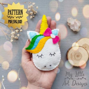 May include: A white felt unicorn head with a yellow horn, rainbow mane, and pink bow. The unicorn has closed eyes and a small black mouth. The image includes the text "PATTERN PDF|SVG|DXF" in a gold starburst.