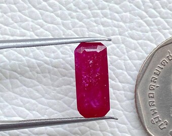 May include: A vibrant, rectangular ruby gemstone held by tweezers, next to a coin. The ruby is a deep, saturated red color with a faceted cut. The coin features a profile of a person and text.