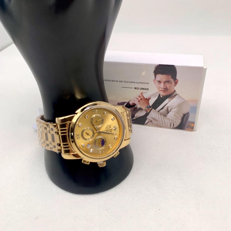 K&ouml;nnte beinhalten: Eine goldfarbene Armbanduhr mit passendem Armband wird auf einem schwarzen St&auml;nder pr&auml;sentiert. Das Zifferblatt der Uhr hat mehrere Hilfszifferbl&auml;tter und eine Datumsanzeige. Im Hintergrund befindet sich eine Schachtel mit einem Foto eines Mannes. Die Uhr ist ein klassisches Accessoire.