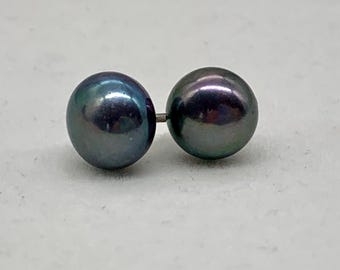 Puede incluir: Un par de pendientes de perlas grises oscuras. Las perlas tienen un brillo iridiscente, que refleja tonos de azul, verde y púrpura. Los pendientes están conectados por un pequeño poste plateado.